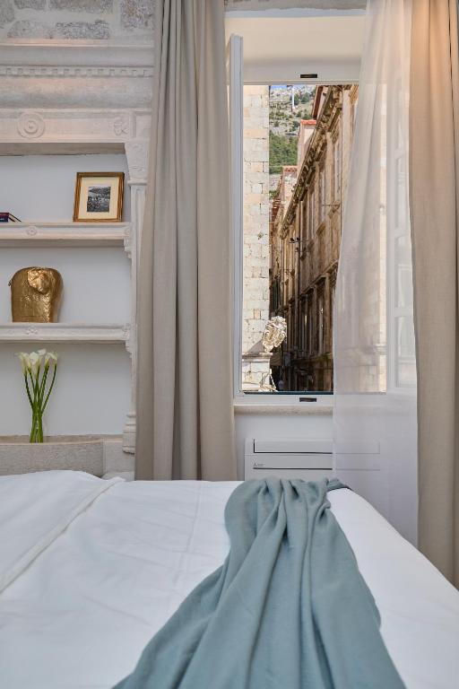 Most Unique Boutique Hotels in Dubrovnik 25 A neatly made bed with white linens and a blue throw faces an open window with beige curtains, offering views typical of boutique hotels in Dubrovnik—a narrow, historic stone street lined with old buildings. Shelves hold a vase with flowers and framed photos.