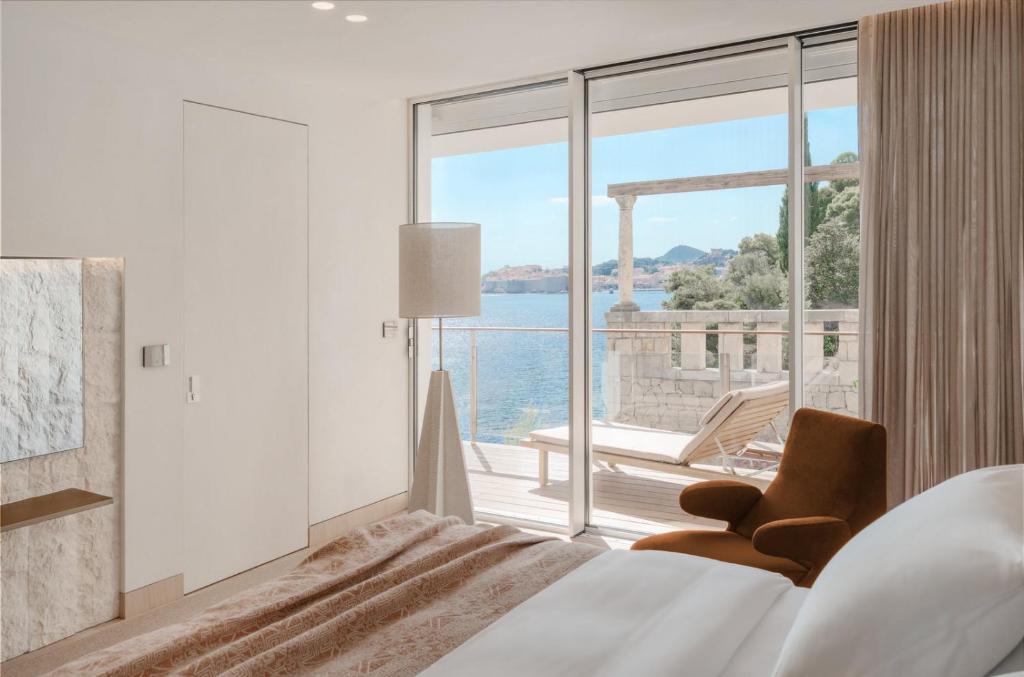 Most Unique Boutique Hotels in Dubrovnik 5 A modern bedroom, inspired by boutique hotels in Dubrovnik, features a large bed, an armchair, and floor-to-ceiling windows opening onto a balcony with lounge chairs—perfect for enjoying scenic views of the water and distant hills.