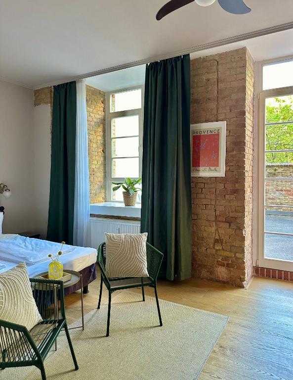 Boutique Hotels in Berlin Kreuzberg 30 A cozy bedroom inspired by boutique hotels in Berlin Kreuzberg, featuring exposed brick walls, green curtains, a large window, a bed, two green chairs with striped cushions, a small table with a yellow vase, and a framed Provence poster on the wall.