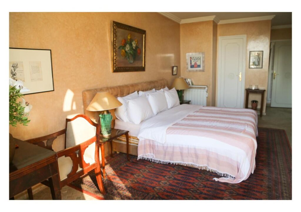9 UNIQUE Riads in Tangier 10 A neatly made bed with white and pink bedding sits in a warmly lit bedroom inspired by riads in Tangier, featuring bedside tables with lamps, framed artwork on beige walls, a wooden chair, and a patterned rug on the floor.