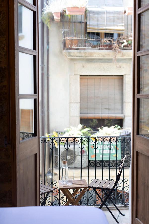 A small balcony with a black wrought iron railing, a folding chair, and a wooden table holding a glass bottle and glass, viewed from inside boutique hotels in Costa Brava through open wooden doors. A neighboring building is visible across the street.