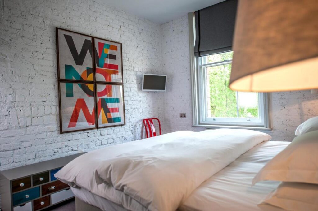 15 Best Boutique Hotels in London, UK 17 A cozy bedroom inspired by the best boutique hotels London offers, featuring white brick walls, a bed with white bedding, a window with a gray shade, a small TV, a red chair, drawers, and wall art that reads WE NOT ME.