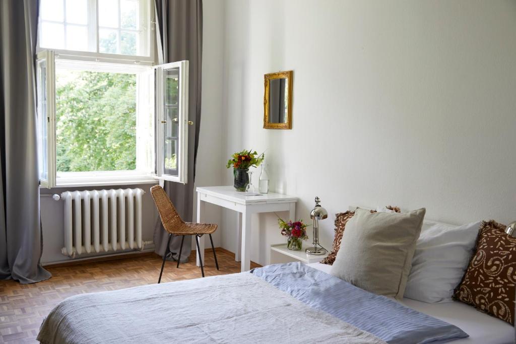 FAIRYTALE Castle Hotels in Bavaria 4 Bright bedroom with a bed, pillows, and blanket, a small desk with flowers, a lamp, and an open window with gray curtains letting in natural light—evoking the charm of castle hotels in Bavaria and views of lush greenery outside.