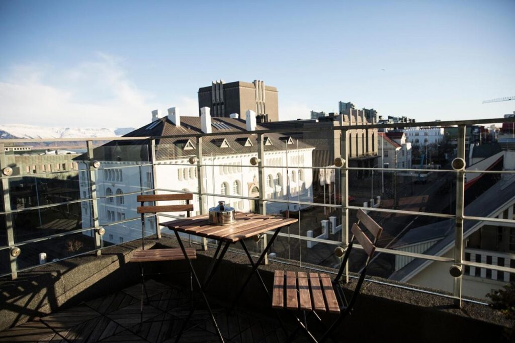 Where to Stay in Reykjavík: Top Guide 7 A small wooden table with two chairs sits on a balcony with glass railing, overlooking city buildings in bright daylight—a perfect spot for those seeking where to stay in Reykjavik, with clear skies and mountains visible in the background.