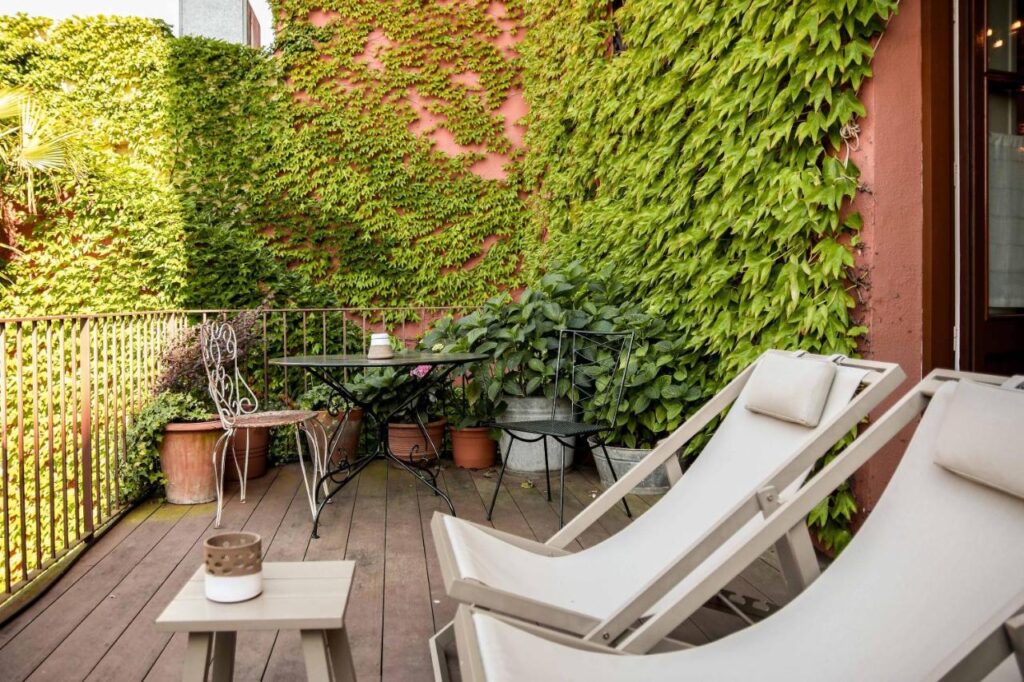 A cozy balcony with two white lounge chairs, a small table, and a black metal table set surrounded by potted plants. The green ivy-covered walls evoke the lush, inviting charm of boutique hotels in Costa Brava.