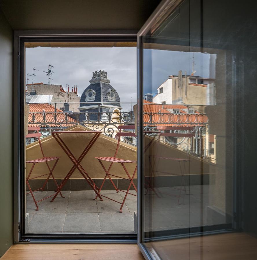 Best Boutique Hotels in San Sebastian 10 A small balcony with a red table and two chairs is seen through an open window, echoing the charm of boutique hotels in San Sebastian, overlooking city rooftops and a decorative dome building under a cloudy sky.
