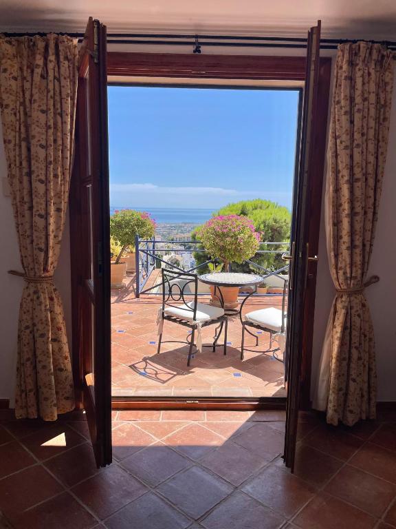 Adults-Only Hotels in Costa del Sol 23 A sunlit terrace with a table and four chairs is seen through open double doors with patterned curtains, reminiscent of the tranquil settings at adult only hotels in Costa del Sol, overlooking greenery and the distant sea under a clear blue sky.