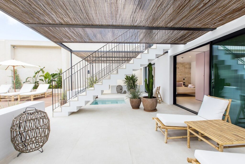Modern outdoor patio with white tile floor, lounge chairs, a bamboo coffee table, potted plants, and a decorative lantern—reminiscent of the serene style found in boutique hotels in Formentera. Sliding glass doors open to an inviting indoor space.