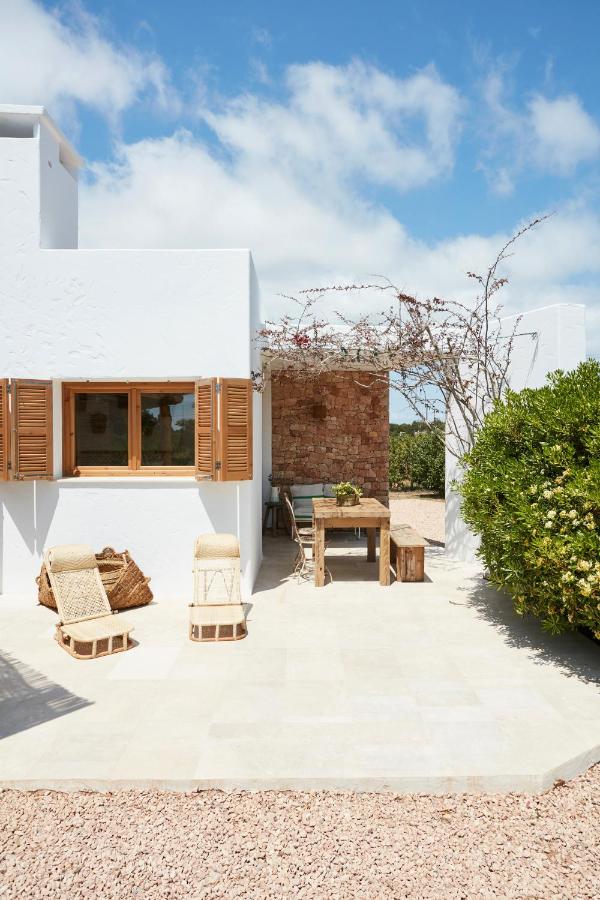 A bright, sunlit patio reminiscent of boutique hotels in Formentera, with white stucco walls, wooden window shutters, wicker chairs, a rustic dining table, and leafy greenery and vines under a blue sky with scattered clouds.