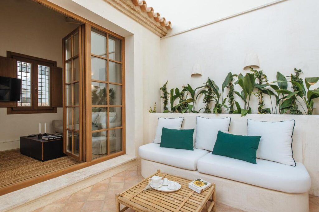 10 UNIQUE Hotels with Pool in Seville 9 A cozy patio in Seville with white walls, a white cushioned sofa with green and white pillows, potted plants along the wall, and a small wooden coffee table—perfect inspiration for guests seeking hotels with pool in Seville.