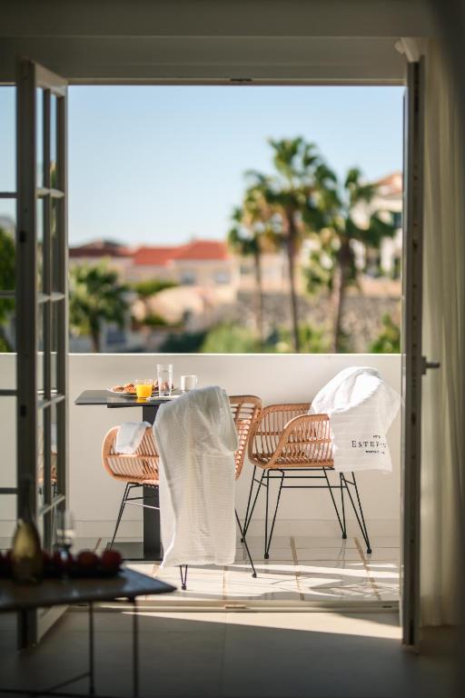 11 UNIQUE Boutique Hotels in Tenerife 30 A sunny balcony in one of the Boutique Hotels in Tenerife features two rattan chairs, a small table set with breakfast, and white robes draped over the chairs. Palm trees and distant buildings can be seen through the open glass doors.