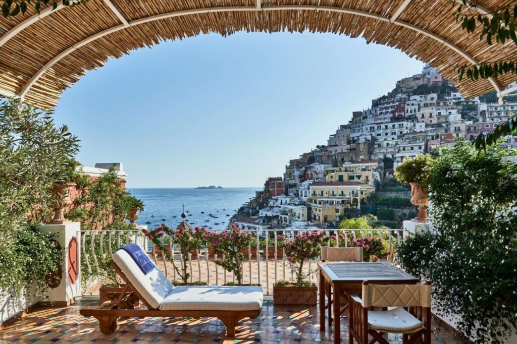 Boutique Hotels on the Amalfi Coast 4 Terrace with lounge chairs and a table overlooks the sea and colorful hillside buildings of Positano, Italy—a scene reminiscent of boutique hotels on the Amalfi Coast—framed by a bamboo pergola and blooming flowers under a clear blue sky.