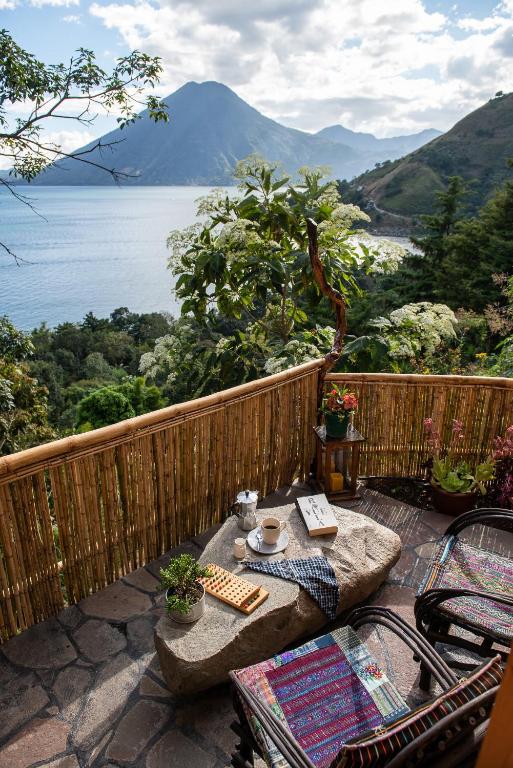 8 UNIQUE Places Where to Stay in Lake Atitlán 22 A cozy stone patio overlooking a lake and mountains, with chairs, a table set for coffee and waffles, books, and colorful woven textiles—an idyllic spot if you’re searching for where to stay in Lake Atitlan, surrounded by lush greenery under a partly cloudy sky.