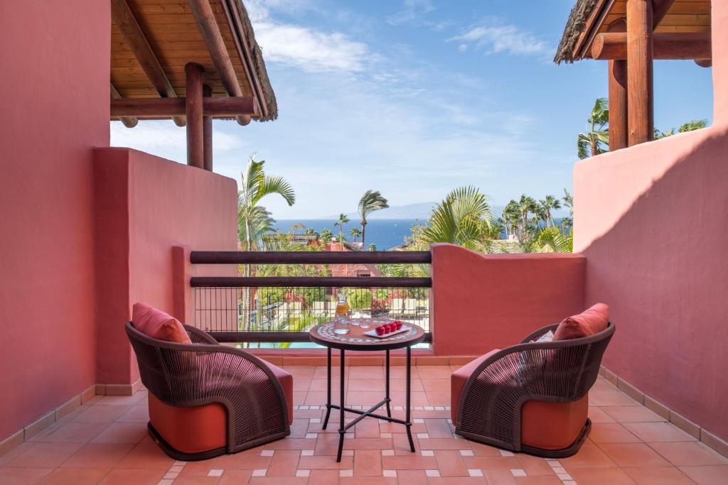 11 UNIQUE Boutique Hotels in Tenerife 47 A terrace at one of the charming Boutique Hotels in Tenerife features two round wicker chairs and a small table overlooking tropical greenery and the ocean, set against salmon-colored walls and tiled flooring under a partly cloudy blue sky.