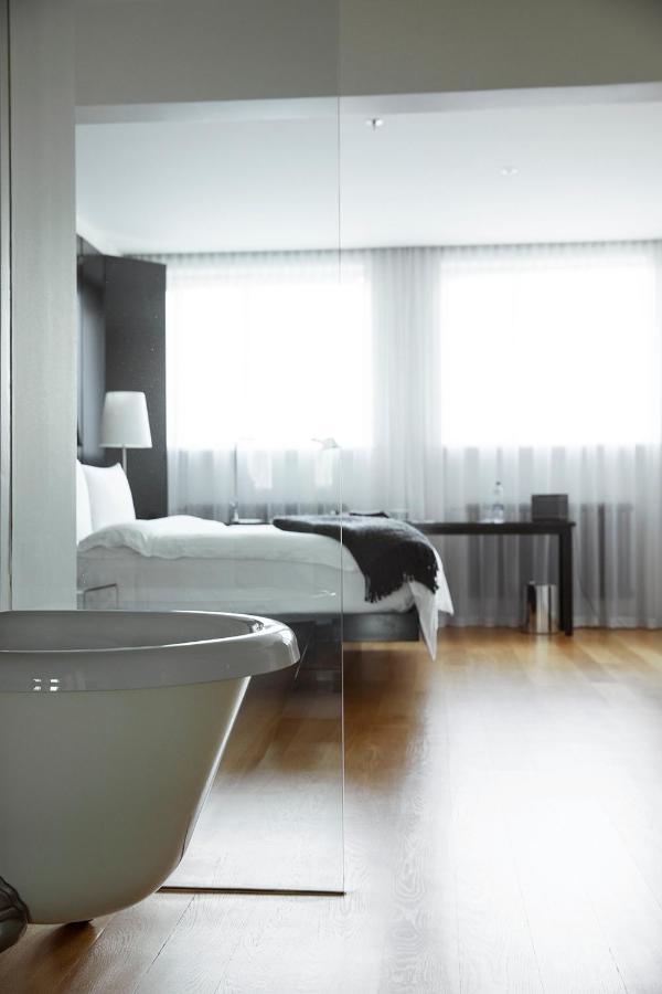 Where to Stay in Reykjavík: Top Guide 6 A modern, minimalistic bedroom—perfect for those seeking where to stay in Reykjavik—with large windows, sheer white curtains, a neatly made bed, side table lamp, desk, and a white freestanding bathtub on wooden flooring.