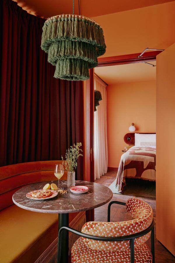 15 Best Boutique Hotels in London, UK 2 A cozy dining nook, reminiscent of the best boutique hotels London offers, features an orange curved bench, round marble table, and patterned chair beside a bedroom. A green tasseled pendant light glows above wine, snacks, and flowers on warm orange-toned walls.