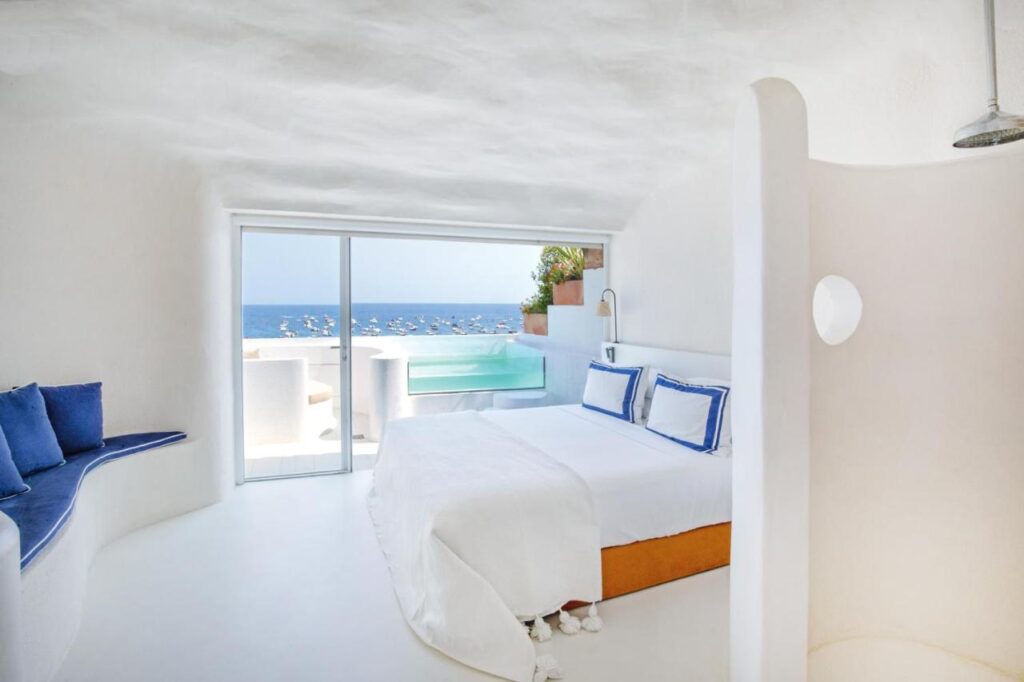 A bright, white, minimalist bedroom with a bed, blue cushions, and sliding glass doors opening to a terrace with a pool and sea view—reminiscent of boutique hotels in Costa Brava, with modern decor creating a coastal, relaxing atmosphere.