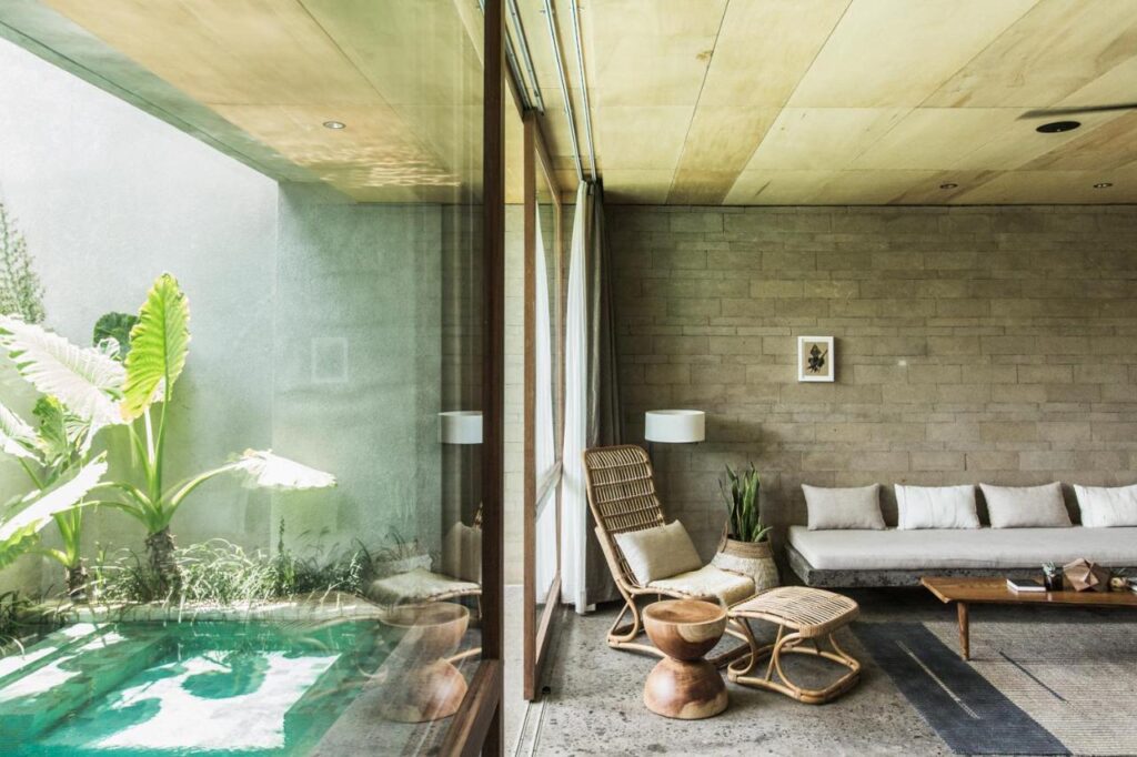14 UNIQUE Boutique Hotels in Canggu, Bali 24 A modern living room with a gray sofa, wicker chairs, plants, and a glass wall overlooking a small pool and lush greenery—reflecting the relaxed elegance found in boutique hotels in Canggu. Natural light and wooden accents complete the minimalist decor.