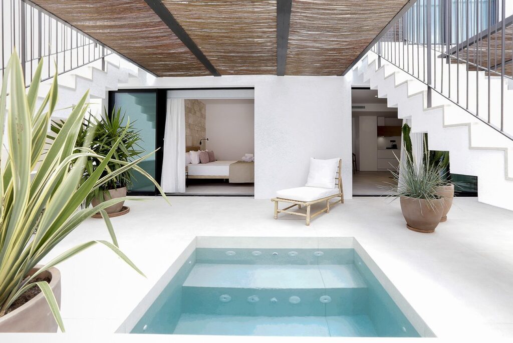 A modern, minimalist patio reminiscent of boutique hotels in Formentera, featuring a small rectangular pool, potted plants, a lounge chair, and two staircases leading up. Sliding doors open to a bedroom and indoor space, with white walls and a slatted roof above.
