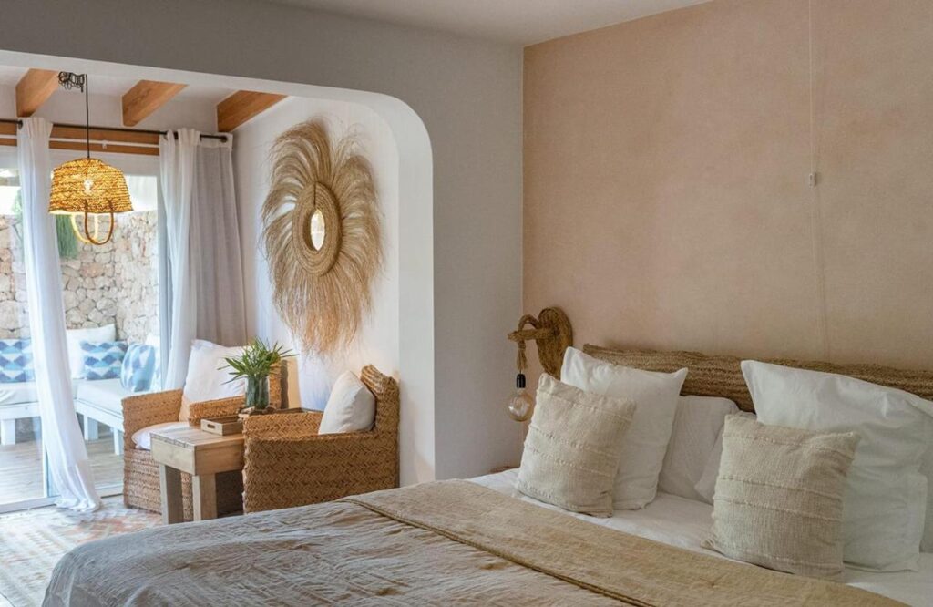 A cozy bedroom with woven decor, a large bed with white and beige pillows, wicker furniture, and a sun-shaped wall hanging—like those found in boutique hotels in Formentera. Natural light pours in from a glass door leading to a bright patio.