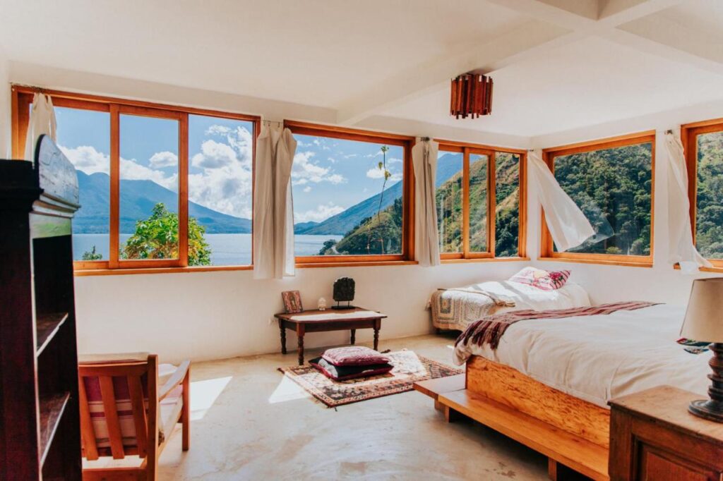 8 UNIQUE Places Where to Stay in Lake Atitlán 10 A bright bedroom with large windows offering scenic views of a lake and surrounding mountains—perfect for those searching where to stay in Lake Atitlan. Wooden furniture, a cozy bed, and sunlight streaming through white curtains create a warm, inviting atmosphere.