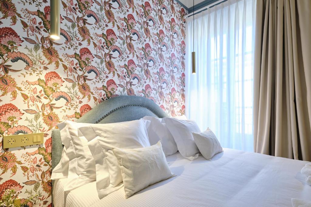 A cozy bedroom with a bed covered in white linens and pillows sits against colorful floral wallpaper, echoing the charm of boutique hotels in Asturias. Soft light streams through sheer curtains, with two modern hanging lamps above creating a bright, inviting feel.