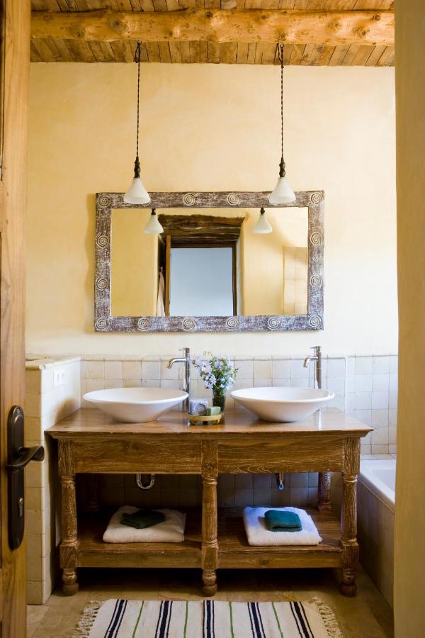 10 AUTHENTIC Finca Hotels in Ibiza 31 Rustic bathroom inspired by finca hotels in Ibiza, featuring a wooden vanity with two vessel sinks, a large decorative mirror, pendant lights, folded towels on the shelf below, a striped rug on the floor, and exposed wood ceiling beams above.