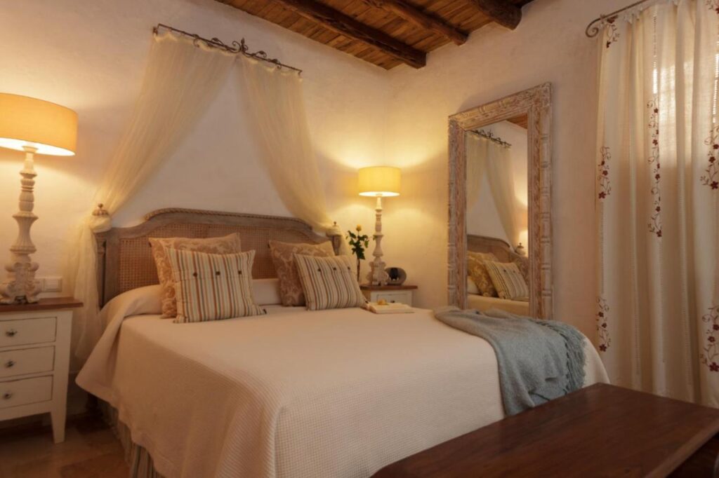 10 AUTHENTIC Finca Hotels in Ibiza 32 A cozy bedroom inspired by finca hotels in Ibiza, with a double bed, striped pillows, and a canopy. Warm lighting from table lamps, an ornate mirror, light curtains, and rustic wooden ceiling beams create an inviting atmosphere.