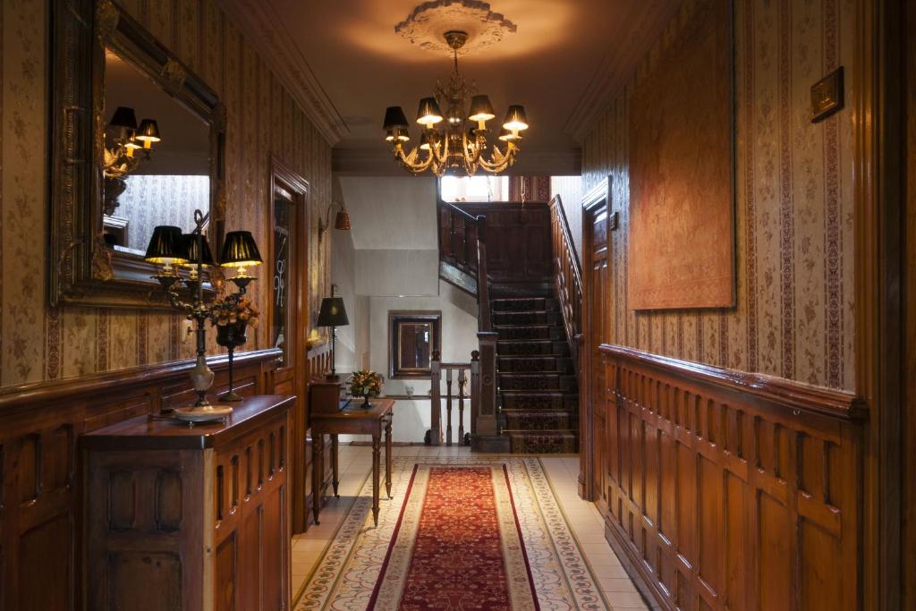A warmly lit hallway with ornate wallpaper, wooden paneling, a red carpet runner, framed artwork, and a chandelier evokes the charm of boutique hotels in Asturias. Furniture and lamps decorate the space, with a staircase leading upstairs in the background.