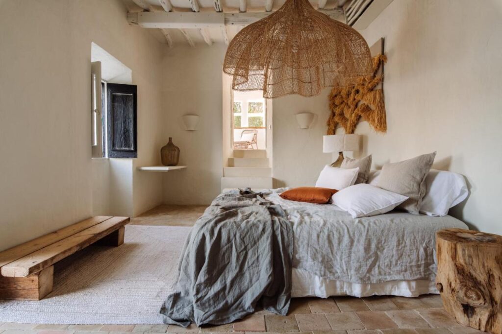 10 AUTHENTIC Finca Hotels in Ibiza 3 A cozy rustic bedroom inspired by finca hotels in Ibiza, featuring a large woven pendant light, a bed with neutral linens and pillows, a wooden bench, earth-toned decor, textured walls, and soft natural light from an open window.