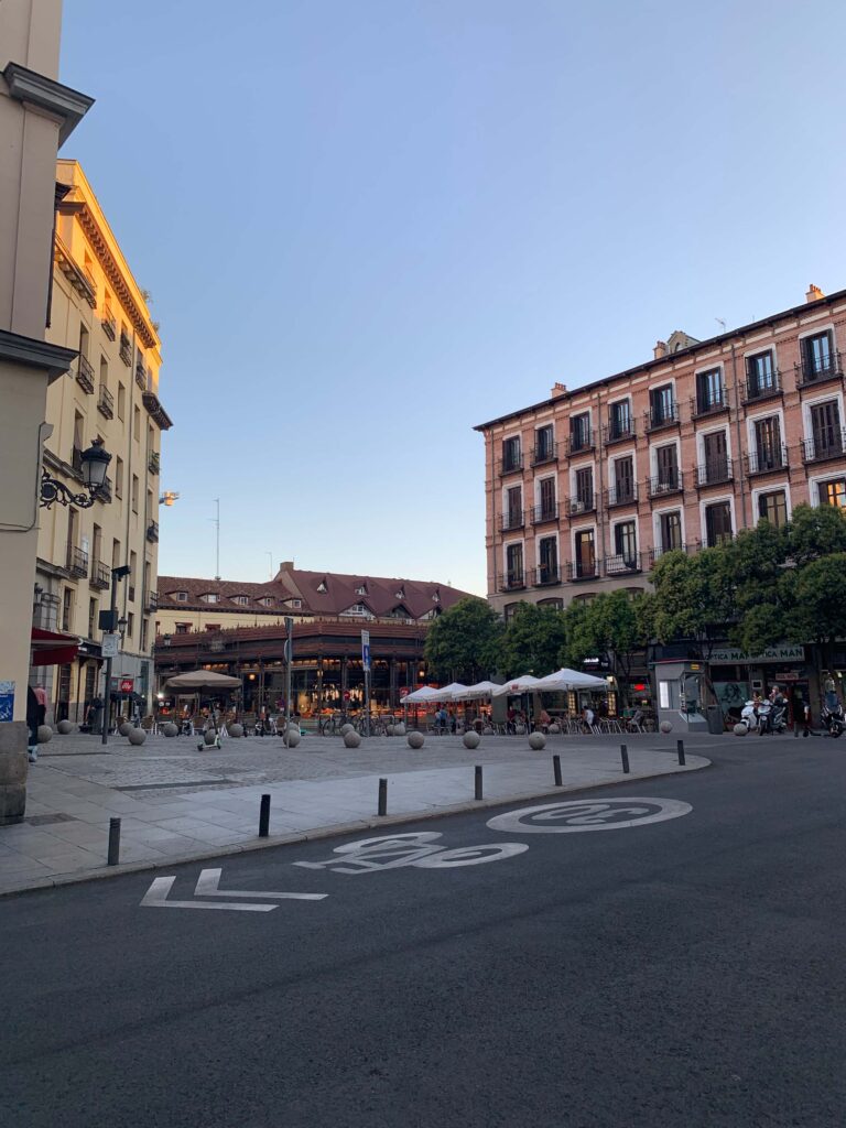 Where to Stay in Madrid: 14 Best Hotels 61 A quiet city square at sunset with outdoor café seating, people dining, trees, and historic buildings—perfect for those searching where to stay in Madrid first time. A bike lane runs along the street in front as the sky remains clear and blue.