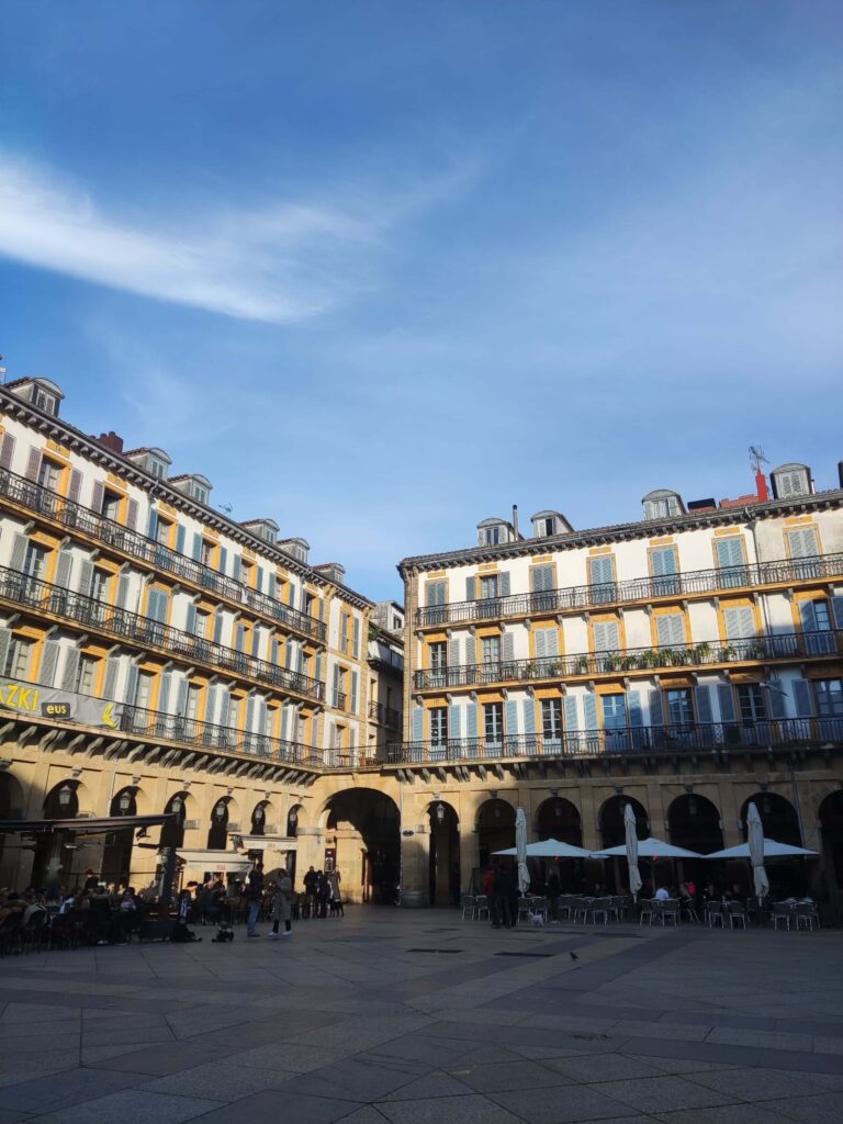 Best Boutique Hotels in San Sebastian 45 Spacious plaza surrounded by historic buildings with rows of windows and balconies, people sitting at outdoor cafes beneath a blue sky, and boutique hotels in San Sebastian just steps away.