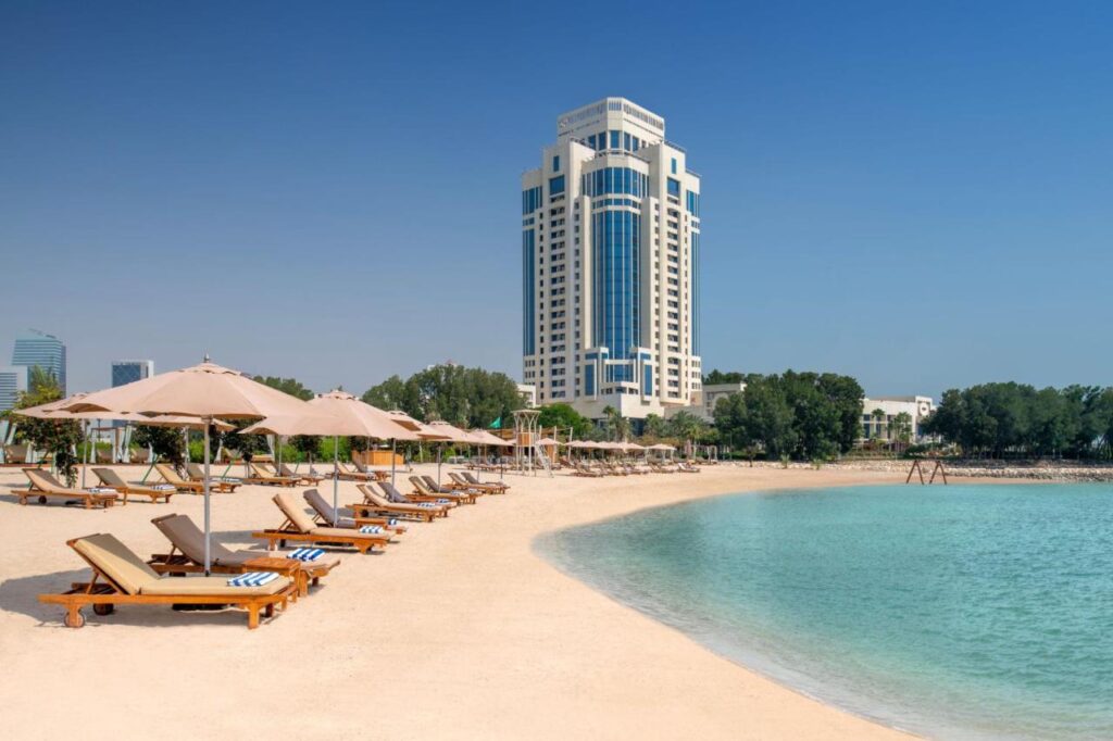 Where to Stay in Doha, Qatar: Transit, City & Beach Hotels 18 A sandy beach with wooden lounge chairs and umbrellas faces calm turquoise water—a perfect spot for travelers searching where to stay in Doha. In the background, a tall modern building rises above green trees under a clear blue sky.