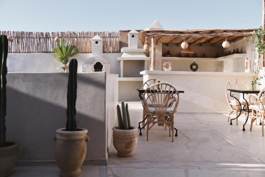 12 Charming Budget Riads in Marrakech 36 Sunny outdoor patio with white stucco walls, potted cacti, wicker chairs, and a rustic bar area—reminiscent of budget riads in Marrakech. Decorative lights hang from a wooden pergola, creating a relaxed, Mediterranean-style atmosphere.