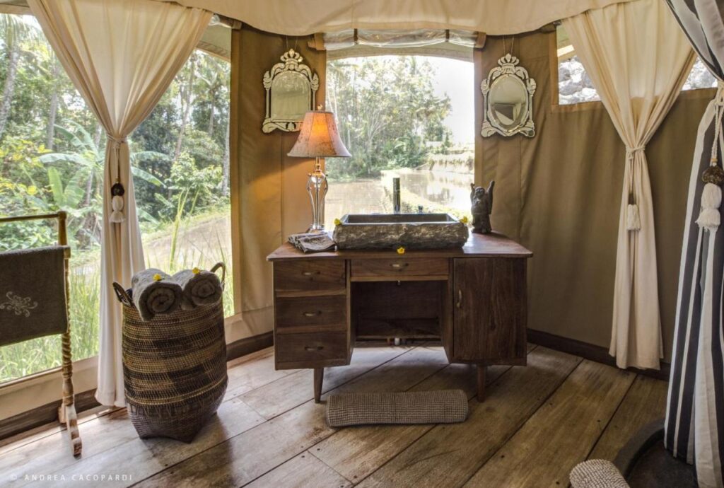 13 Most UNIQUE Places to Stay in Ubud 33 A rustic wooden vanity with a stone sink and lit lamp sits inside a tent with large windows, two ornate mirrors, and rolled towels in a basket—lush greenery is visible outside, offering one of the most unique places to stay in Ubud.