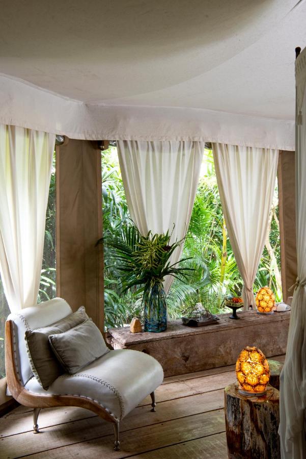 13 Most UNIQUE Places to Stay in Ubud 31 A cozy lounge chair sits by large windows with white curtains, overlooking lush green foliage. A vase with tropical leaves and glowing lamps create a relaxing vibe—perfect for those seeking unique places to stay in Ubud.