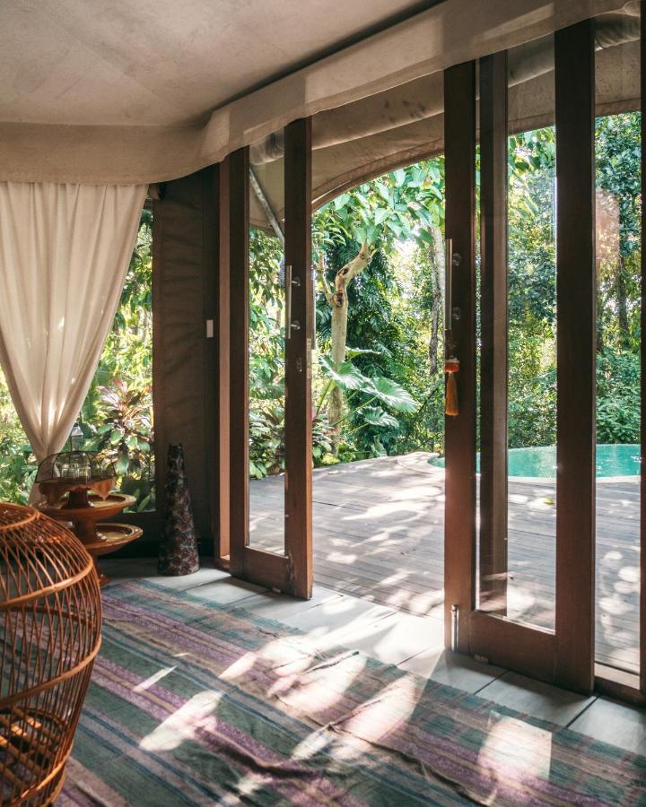 13 Most UNIQUE Places to Stay in Ubud 32 Sunlight streams through glass doors onto a striped rug inside a cozy room with natural wood accents, wicker furniture, and light curtains—one of the unique places to stay in Ubud, opening to a wooden deck and lush green jungle outside.
