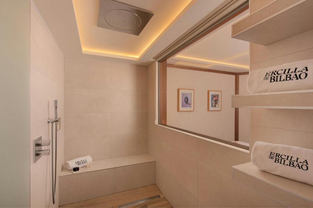 8 Unique Boutique Hotels in Bilbao Spain 16 A modern, beige-tiled shower in one of the Boutique Hotels in Bilbao features a rain showerhead, a built-in bench, and shelves with folded towels labeled Ercilla de Bilbao. A large window reveals a room adorned with framed art.