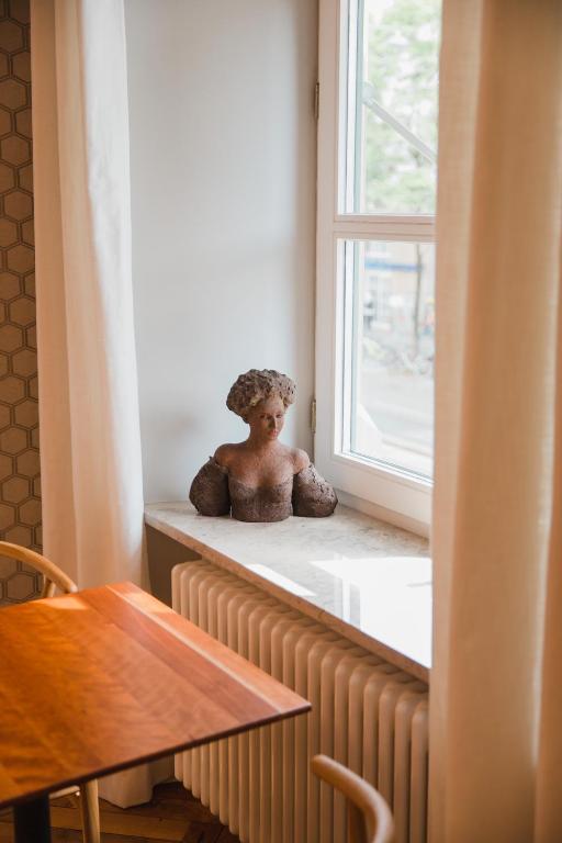 Authentic Boutique Hotels in Munich 25 A small bust sculpture sits on a windowsill next to a white radiator, reminiscent of elegant boutique hotels in Munich. Light streams through the window, partially covered by light curtains. Part of a wooden table and chair appear in the foreground.