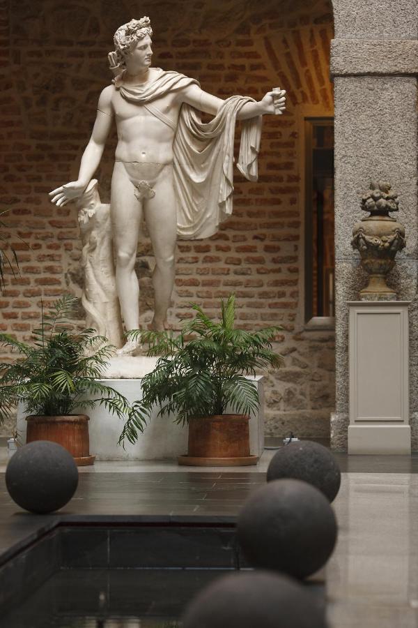 25 Weekend Trips from Madrid Max. 2h Away 92 A marble statue of a nude male figure stands on a pedestal surrounded by potted plants, evoking the charm of hidden courtyards often discovered on weekend trips from Madrid, with round black stones accenting a water feature in the foreground.