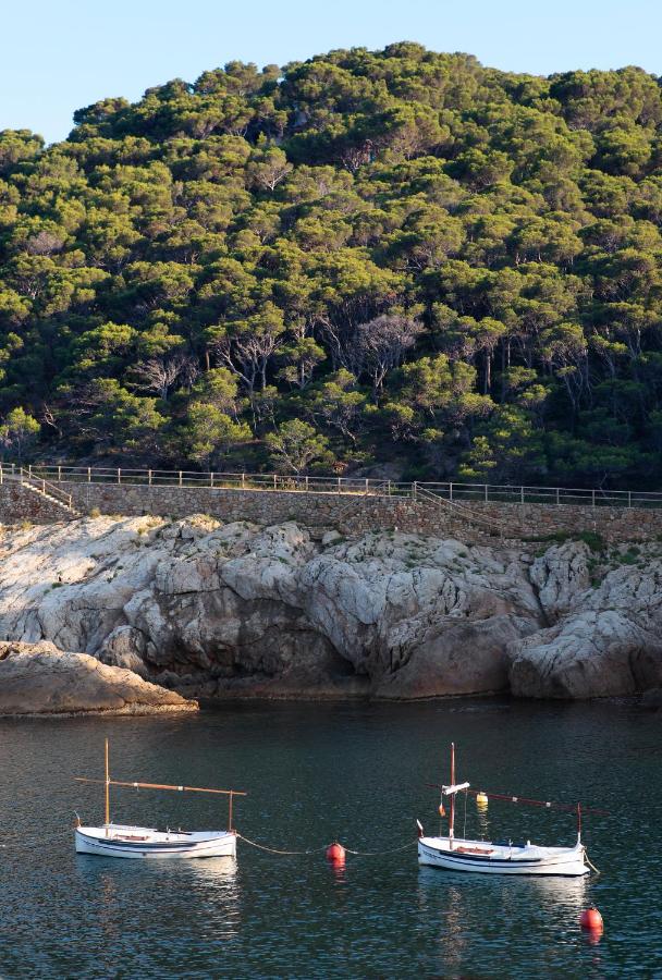 Two small white boats float on calm water near a rocky shoreline, framed by a dense green forest and wooden fence—typical of the tranquil scenery found near boutique hotels in Costa Brava. Red and orange buoys surround the peaceful boats.