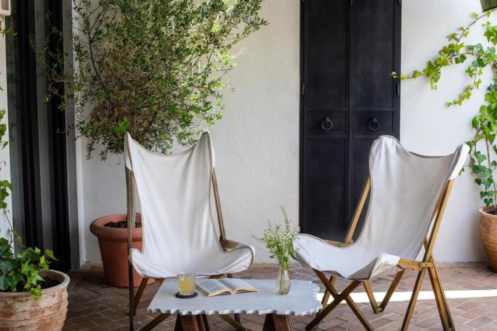 Where to Stay in Madrid: 14 Best Hotels 33 Two white canvas lounge chairs face a small table with a book, flowers, and juice on a patio—an inviting spot when considering where to stay in Madrid first time visitors will love, framed by potted plants and a black door on a white wall.