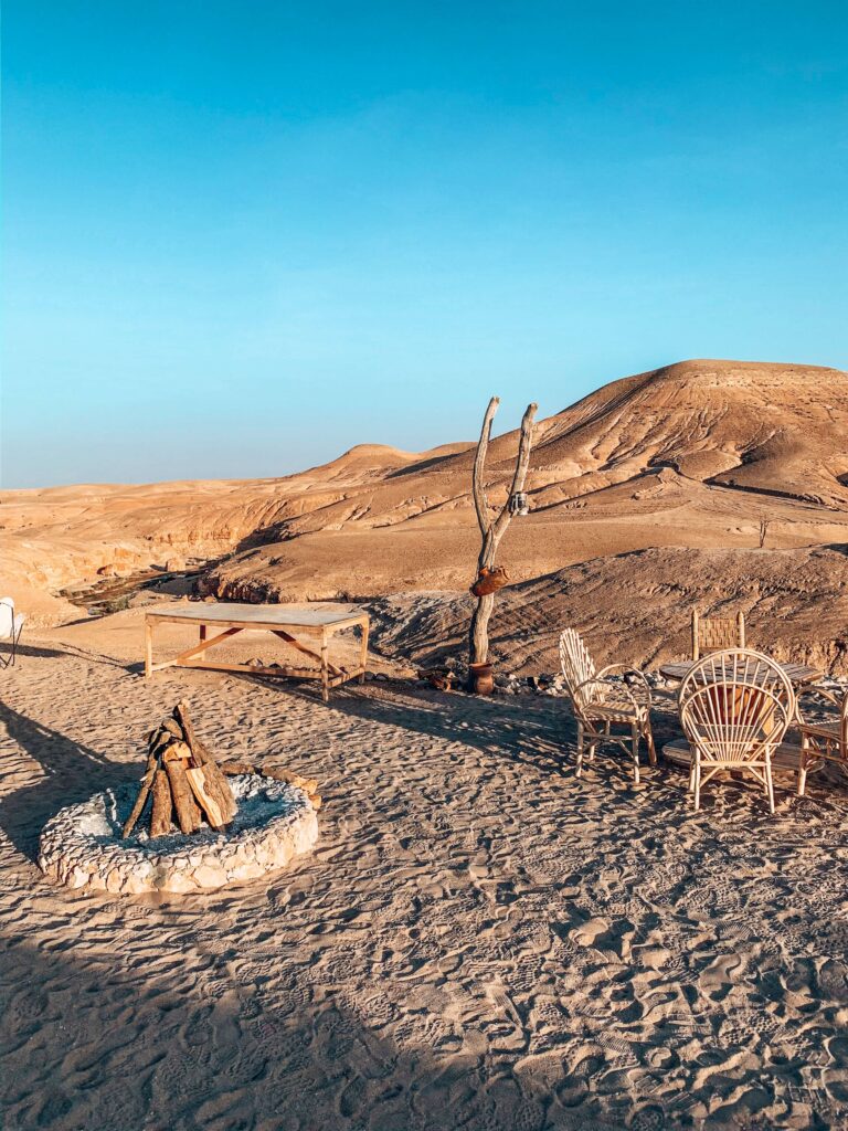 8 SPECTACULAR Agafay Desert Camps 45 A desert scene with sandy hills, wicker chairs around a stone fire pit, a wooden table, and a leafless tree under a clear blue sky, reminiscent of tranquil Agafay Desert camps.