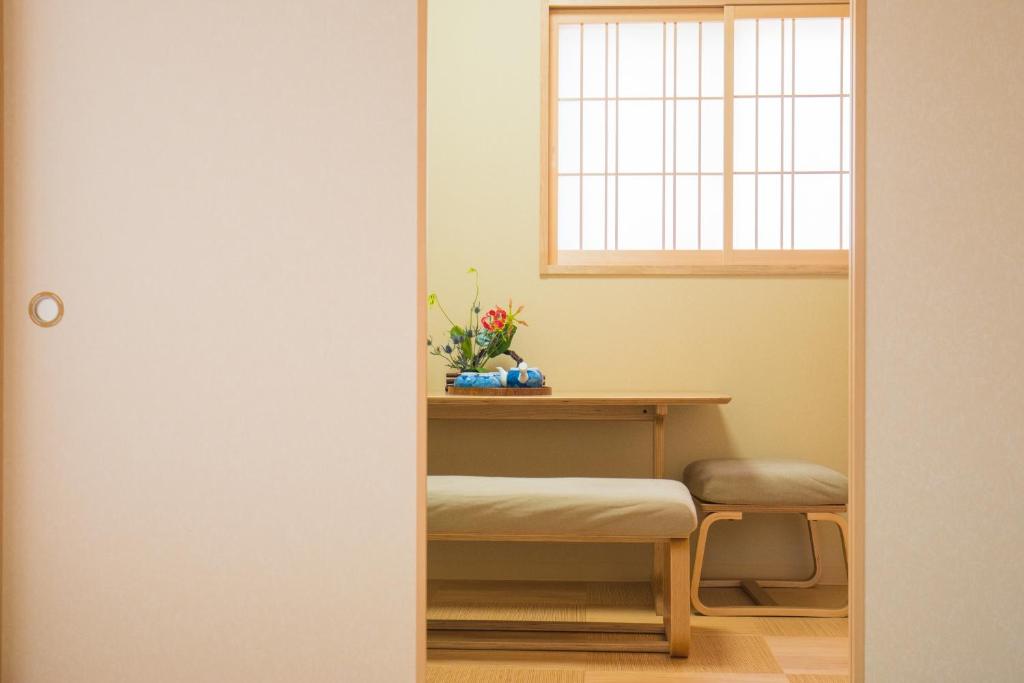 8 BEST Ryokans in Osaka 12 A minimalist Japanese-style room inspired by ryokans in Osaka, with light wooden furniture, a low table, two cushioned benches, a flower arrangement, and a shoji window letting in soft natural light.