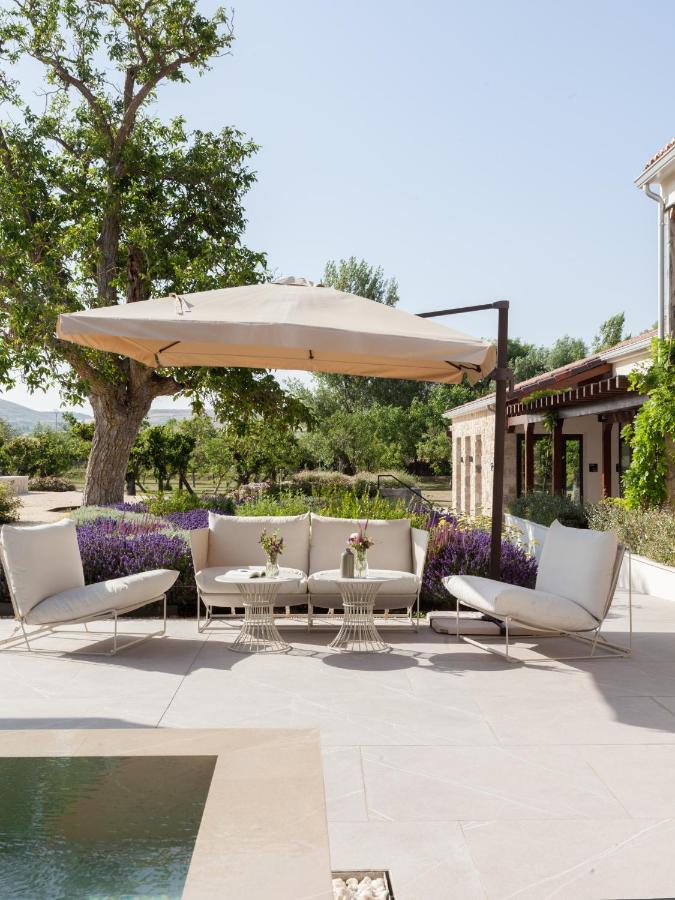 25 Weekend Trips from Madrid Max. 2h Away 65 A modern outdoor patio with white cushioned seating, small round tables, and a large beige umbrella—perfect for relaxing during weekend trips from Madrid. Blooming lavender, trees, and a rustic building complete the tranquil scene.