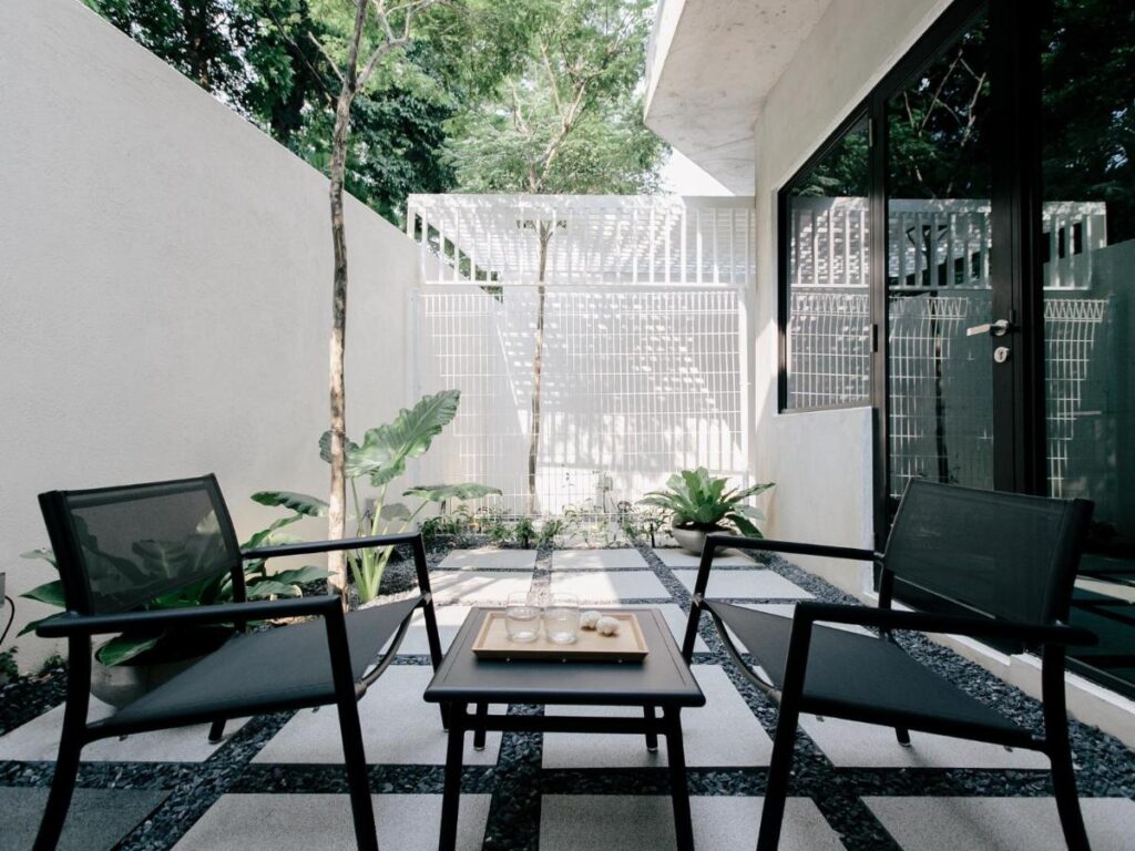 13 Most Unique Hotels in Singapore for an Incredible Holiday 13 A modern outdoor patio at one of the unique hotels in Singapore, with two black mesh chairs and a small wooden table on tiled flooring, surrounded by lush plants, white walls, and large glass windows.