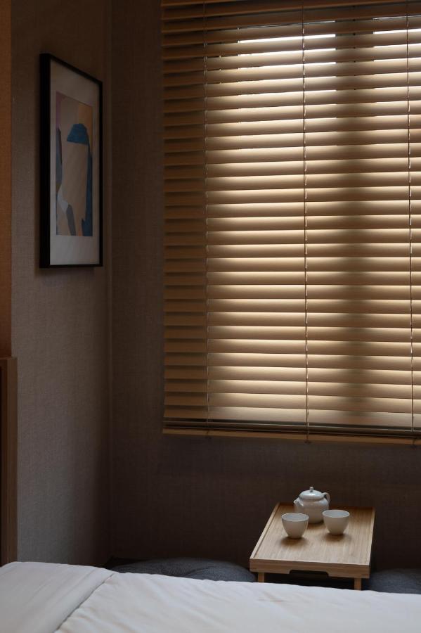13 Most Unique Hotels in Singapore for an Incredible Holiday 26 A cozy bedroom corner in one of the unique hotels in Singapore, featuring closed beige blinds, soft lighting, white-sheeted bed, a wooden tray with a teapot and cups, and framed abstract artwork on the wall.