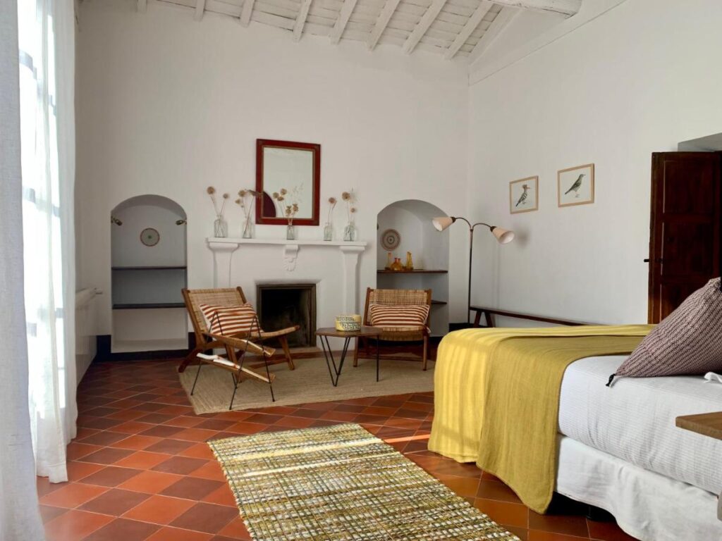 25 Weekend Trips from Madrid Max. 2h Away 13 Bright, cozy bedroom with terracotta tile floor—perfect for weekend trips from Madrid. Features a double bed with yellow blanket, rattan chairs by a white fireplace, minimalist decor, and framed art. Natural light fills the inviting space.