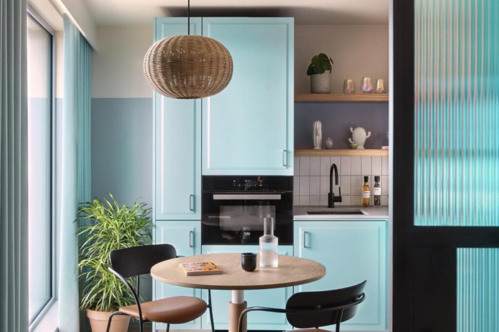 15 Best Boutique Hotels in London, UK 27 A modern kitchen with light blue cabinets, a round wooden table with two chairs, a black faucet, open shelves, and a woven pendant lamp—evoking the stylish charm of the best boutique hotels London has to offer. Sunlight streams in through large windows.