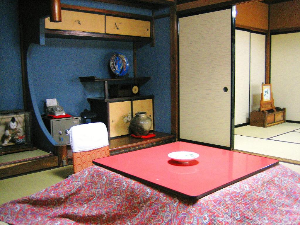 Looking for the Best Ryokan in Takayama? 9 Traditional Japanese tatami room inspired by the Best Ryokan in Takayama, with a red kotatsu table, patterned cloth, sliding shoji doors, wooden shelves, pottery jar, decorative items, and blue and cream-colored walls.